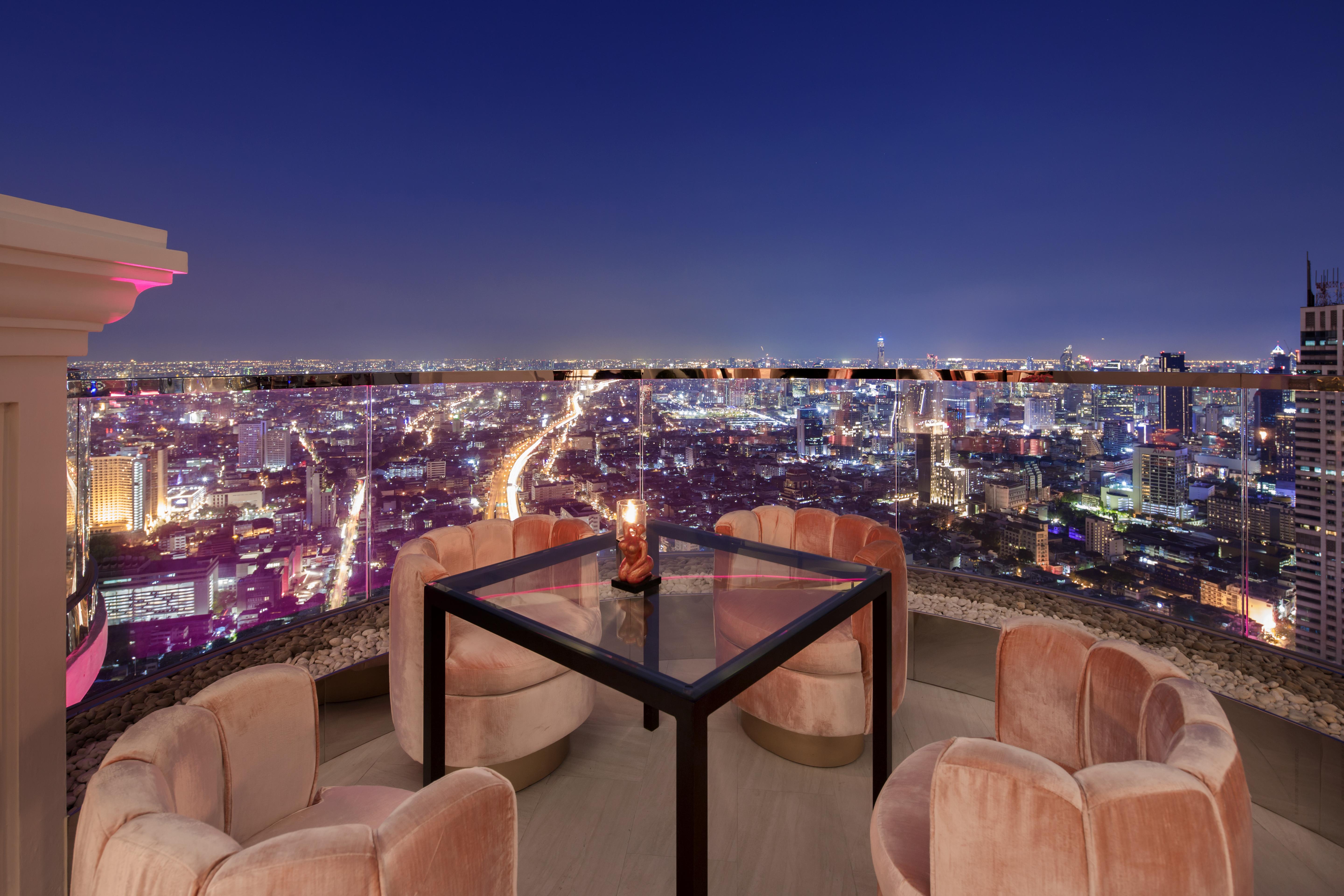 Lebua At State Tower Hotel Банкок Екстериор снимка The photo depicts a stylish outdoor seating area on a rooftop, featuring a square glass table surrounded by four plush, circular chairs. The scene is set against a stunning nighttime city skyline, with a multitude of twinkling lights visible in the b